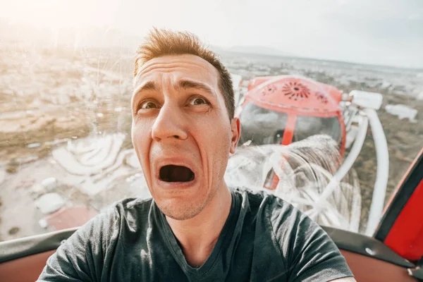 A guy with acrophobia or fear of heights screams with funny emotions on his face from the view from a high Ferris wheel in an amusement park