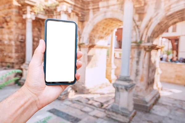 Hand with blank white screen of smartphone against background of Hadrian Arch in Antalya old town. Travel application and digital guide concept