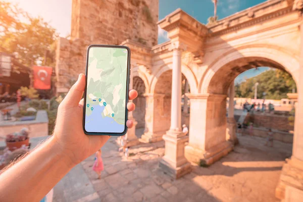 Hand with screen of smartphone with working maps and geolocation with checkpoints against background of Hadrian Arch in Antalya old town. Travel applications and digital guide concept