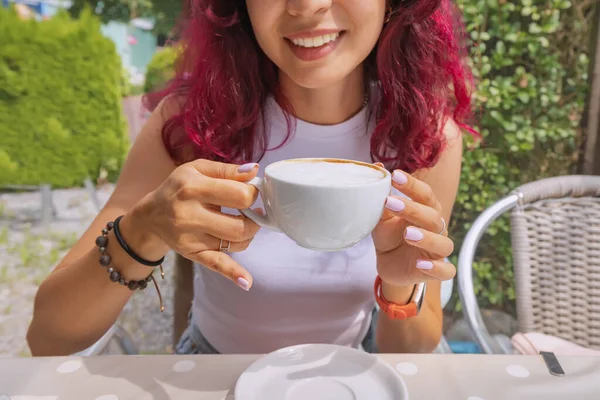 The girl smiles with snow-white teeth and drinks cappuccino coffee in a cafe