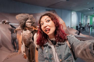 22 Temmuz 2022, Neanderthal Müzesi, Almanya: Müzedeki tarih öncesi atalarının balmumu figürleriyle selfie çeken komik modern homo sapiens kızı. Antropoloji bilim konsepti