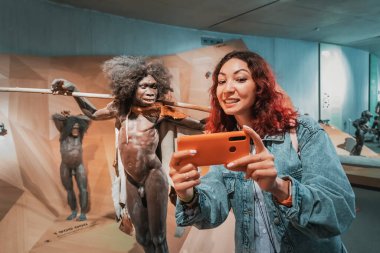 22 Temmuz 2022, Neanderthal Müzesi, Almanya: Müzedeki tarih öncesi atalarının balmumu figürleriyle selfie çeken komik modern homo sapiens kızı. Antropoloji bilim konsepti