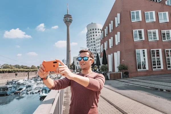 Happy man travel blogger takes selfie pictures on smartphone of the famous Dusseldorf TV tower from the Media Harbor canal in the post industrial district. Travel and sightseeing locations