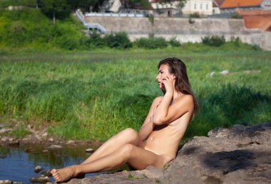 Genç çıplak kadın eski bir taşra kasabasında nehrin kıyısındaki bir taşta güneşleniyor.