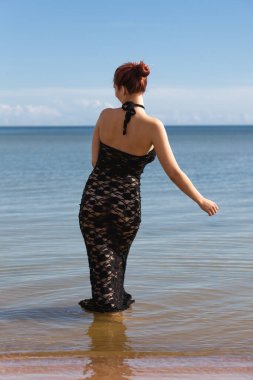 Siyah dantel elbiseli genç, kızıl saçlı kadın sahilde çıplak poz veriyor.