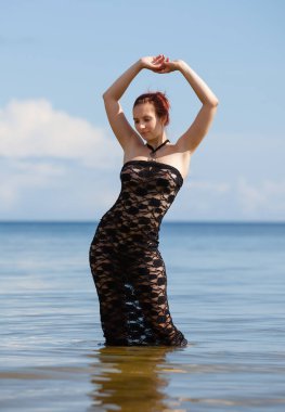 Siyah dantel elbiseli genç, kızıl saçlı kadın sahilde çıplak poz veriyor.