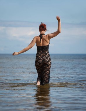 Siyah dantel elbiseli genç, kızıl saçlı kadın sahilde çıplak poz veriyor.