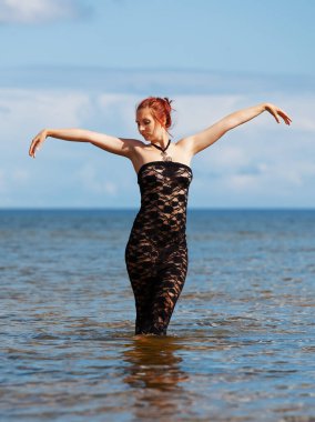 Siyah dantel elbiseli genç, kızıl saçlı kadın sahilde çıplak poz veriyor.