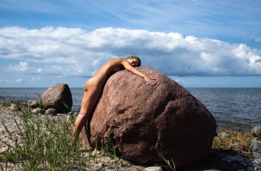 Genç, narin bir kadın doğanın tadını çıkarıyor. Deniz kıyısında çıplak bir kadın güneşli bir günde büyük bir taşın yanında poz veriyor.