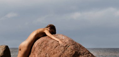 Genç, narin bir kadın doğanın tadını çıkarıyor. Deniz kıyısında çıplak bir kadın güneşli bir günde büyük bir taşın yanında poz veriyor.