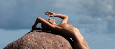 Genç, narin bir kadın doğanın tadını çıkarıyor. Deniz kıyısında çıplak bir kadın güneşli bir günde büyük bir taşın yanında poz veriyor.