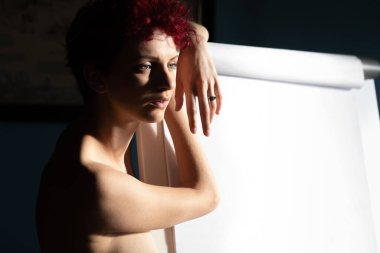 İç taraftaki çıplak kızıl saçlı kadın doğal ışıkla aydınlatılmış pencerenin yanında.