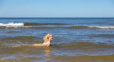 Yaşam tarzı ve çıplaklık kavramı. Tamamen çıplak, güzel bir sarışın kadın güneşli bir günde deniz kıyısında doğanın tadını çıkarıyor..