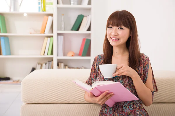 Happy asian girl reading on sofa while enjoying coffee — Zdjęcie stockowe