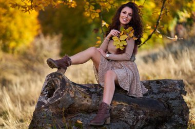 Güzel, zarif genç bir kadın, yaşlı bir ağacın gövdesinin altında, sonbaharın canlı renklerinde poz veriyor.