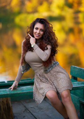 Güzel bir manken, göle giden bir iskelede oturuyor. Etrafı renkli ormanlarla çevrili. Suyu yansıtıyor.