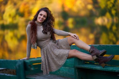 Güzel bir manken, göle giden bir iskelede oturuyor. Etrafı renkli ormanlarla çevrili. Suyu yansıtıyor.