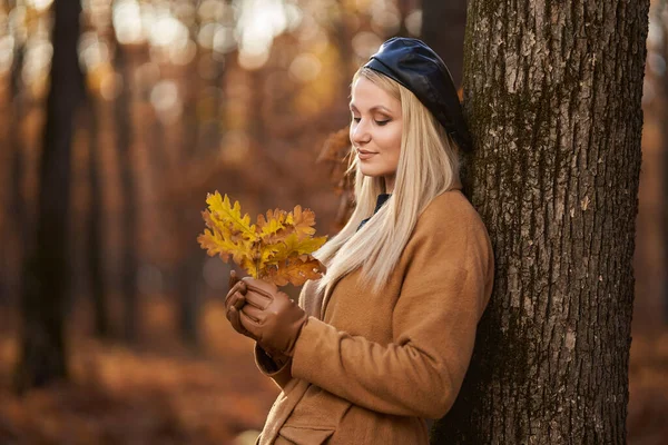 在生机勃勃的橡木森林里 一个身穿大衣戴着帽子的金发年轻女子的自拍直率画像 — 图库照片