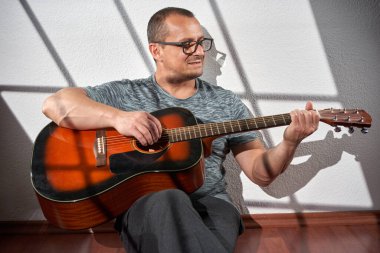 Adam akustik gitar çalıyor, yerde oturuyor, pencereden yansıyan ışıkla birlikte.