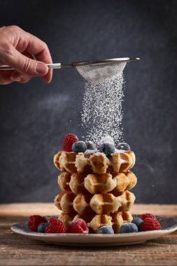Bir kadının eli, üzerinde taze böğürtlen olan waffle yığınına pudra şekeri serpiyor.