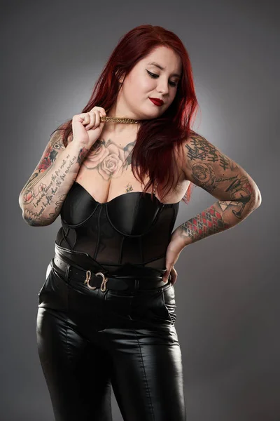 Glamour plus size model covered in tattoos in lingerie and leather on gray background