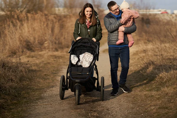 年轻的家庭带着他们的小女儿带着婴儿车在住宅区走过公园 — 图库照片
