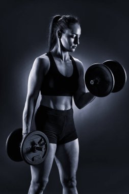 Güçlü, güzel bir kadın halterlerle bisepsi kıvırıyor, ayakta duruyor.