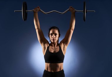 Young athletic woman doing shoulder workout with barbell on blue background
