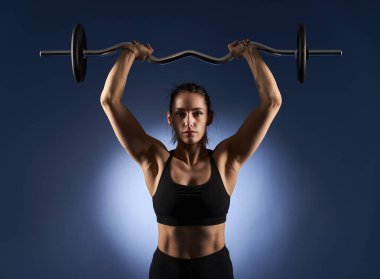 Young athletic woman doing shoulder workout with barbell on blue background