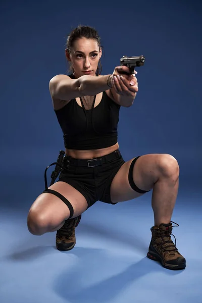 黒のショートパンツとトップのアスレチック冒険家の若い女性 銃と戦術的なナイフで 青の背景に — ストック写真