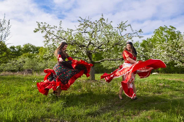 Geleneksel çingene kostümlü iki genç kadın çiçek açan bir elma bahçesinde dans ediyor.