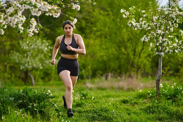 果樹園で草の上を走る美しいプラスサイズのランナーインドの女性 — ストック写真