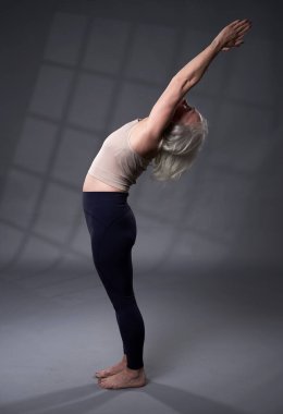 Beyaz saçlı, yoga eğitimi almış yaşlı bir kadın, gri arka planda stüdyo çekimi.