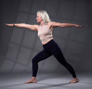 Beyaz saçlı, yoga eğitimi almış yaşlı bir kadın, gri arka planda stüdyo çekimi.
