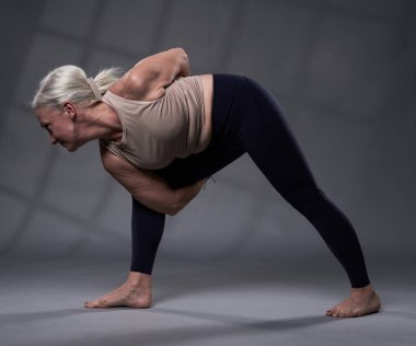 Beyaz saçlı, yoga eğitimi almış yaşlı bir kadın, gri arka planda stüdyo çekimi.