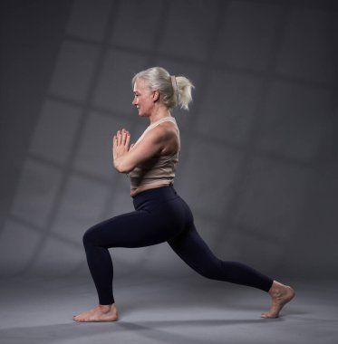 Beyaz saçlı, yoga eğitimi almış yaşlı bir kadın, gri arka planda stüdyo çekimi.