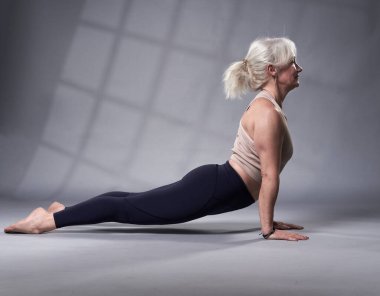 Beyaz saçlı, yoga eğitimi almış yaşlı bir kadın, gri arka planda stüdyo çekimi.