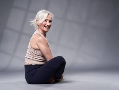 Beyaz saçlı, yoga eğitimi almış yaşlı bir kadın, gri arka planda stüdyo çekimi.