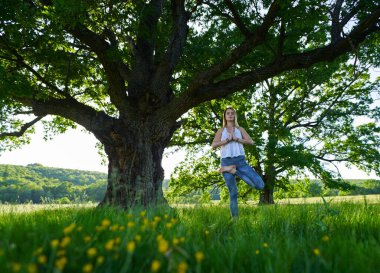 Yüzüncü yıl meşe ağaçlarıyla dolu bir ormanda yoga ve meditasyon yapan genç bir kadın.
