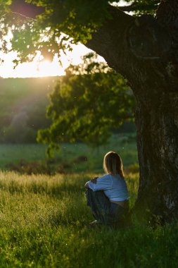 Gün batımında kocaman bir meşe ağacının yanında poz veren çekici sarışın kadın.