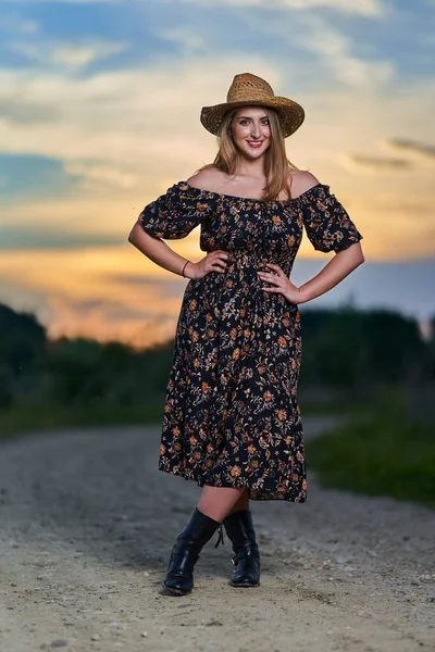 プラスサイズ魅力的な農家の女性で花のドレスと麦藁帽子で田舎道で日没 — ストック写真