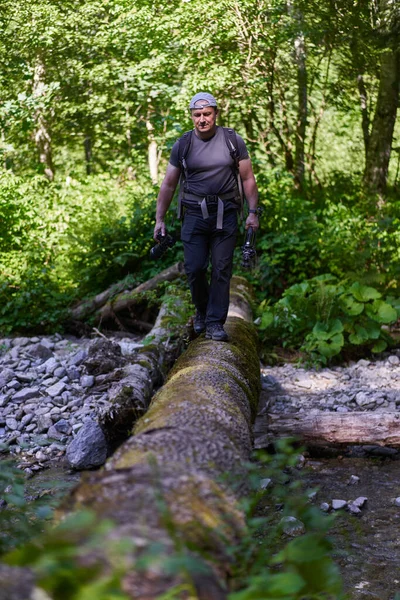Male nature photographer with heavy backpack crossing a river on a fallen tree