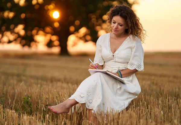 Woman writer in white dress with her notebook in a harvested field with a huge oak in the background at sunset