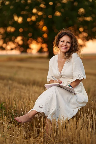 Woman writer in white dress with her notebook in a harvested field with a huge oak in the background at sunset