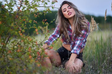 Ekose gömlekli ve kısa pantolonlu güzel bir kadın gün batımında kırsalda, çalıların yanında.