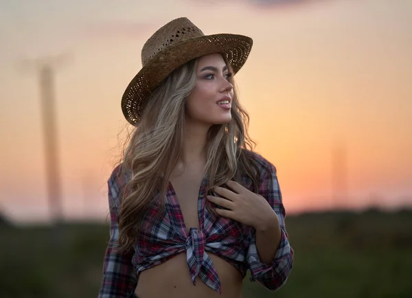 Beautiful cowgirl in hat, plaid shirt and short jeans at sunset in a countryside field