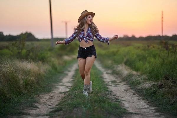 Beautiful cowgirl in hat, plaid shirt and short jeans at sunset in a countryside field