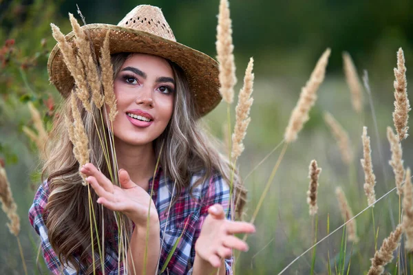 Beautiful cowgirl in hat, plaid shirt and short jeans at sunset in a countryside field