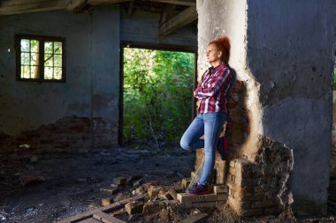 Kıvırcık saçlı, ekose gömlekli ve kot pantolonlu bir kadın harap bir ahırda ve evde.