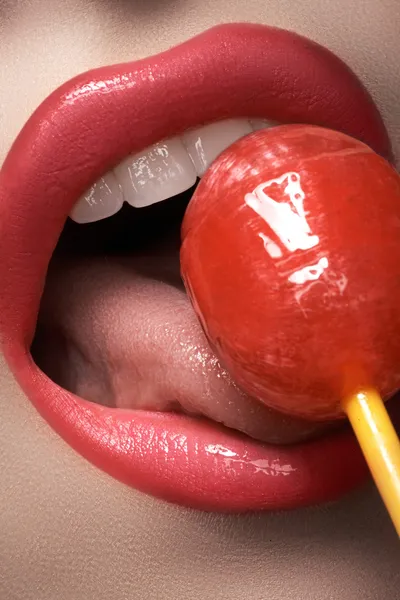 Macro atirar glamour com lábios de mulher sexy com um bombom doce. Linda maquiagem de moda brilhante. Menina lambendo um chupa-chupa de morango. Dentes saudáveis, sorriso engraçado — Fotografia de Stock